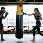 Side view of determined fit young female boxer in sportswear and gloves punching bag during workout with male personal trainer standing in stance in modern gym