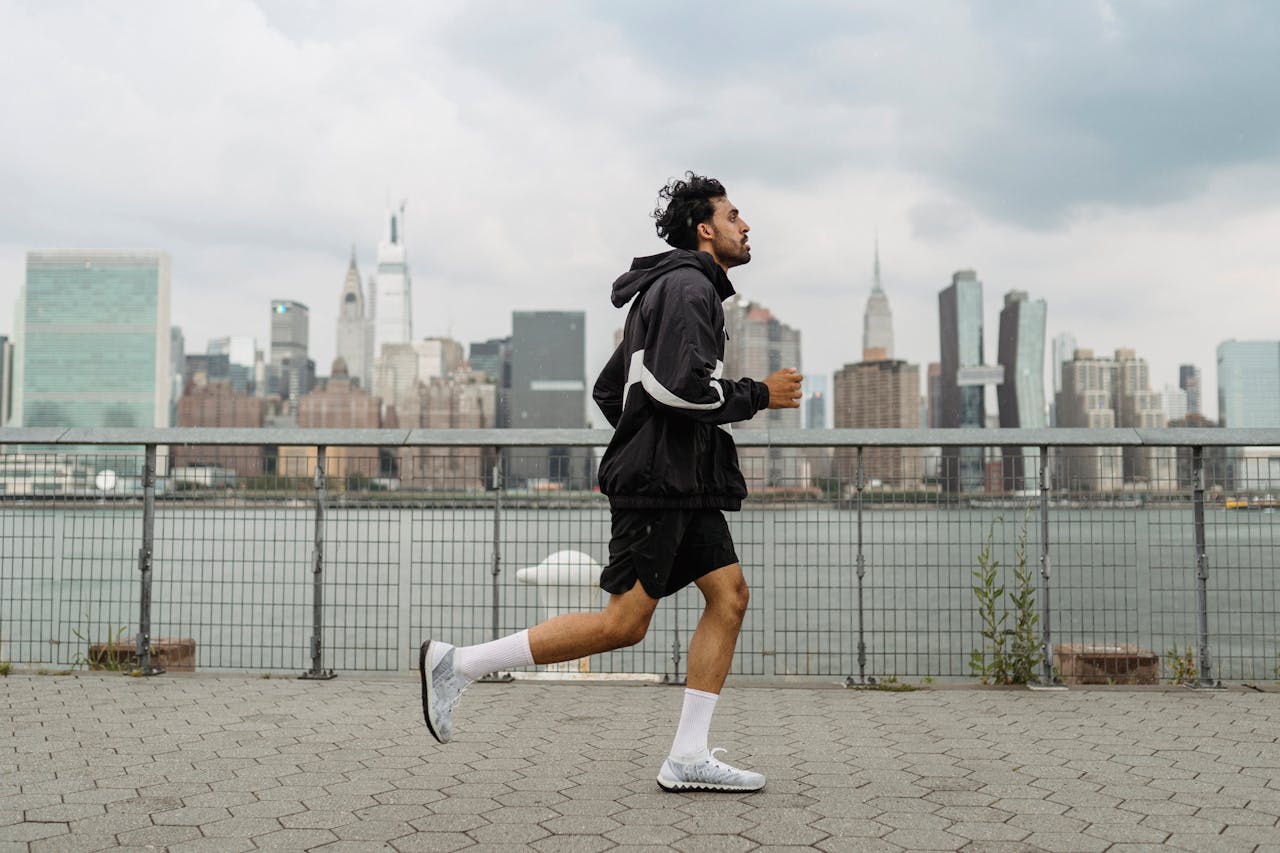 services-09 Adult man jogging along a waterfront with the New York City skyline in the background, exuding a vibrant urban lifestyle.